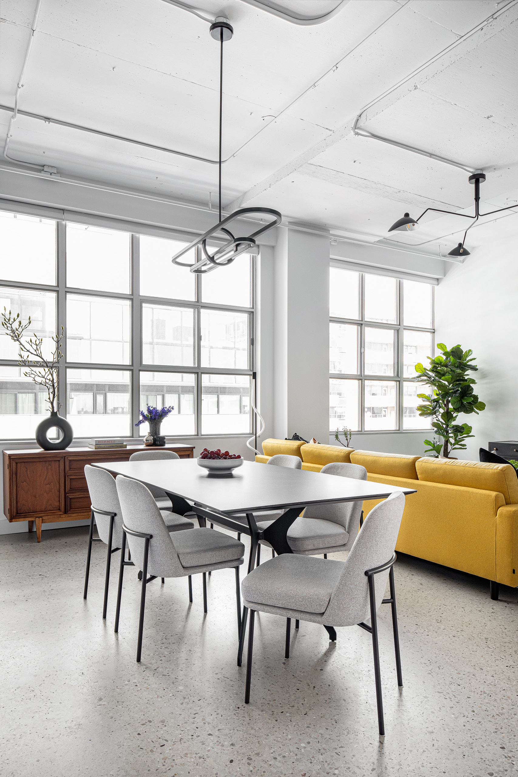 Bright Eglinton loft living space featuring expansive warehouse windows, a black ceramic dining table, and a striking large yellow sectional sofa.