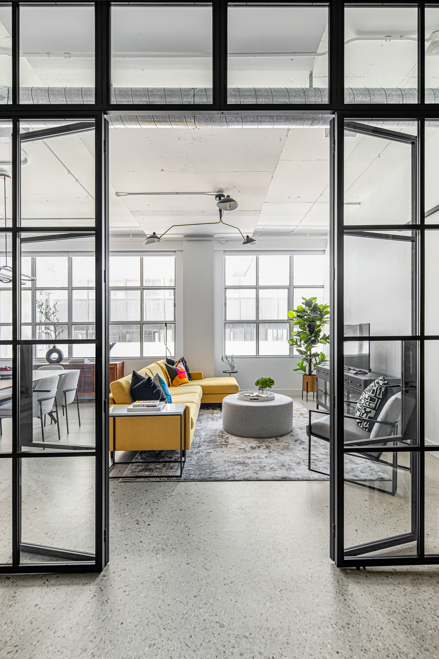 Looking through open custom black metal and glass library doors into a bright loft living room featuring a yellow sectional and exposed concrete ceilings.