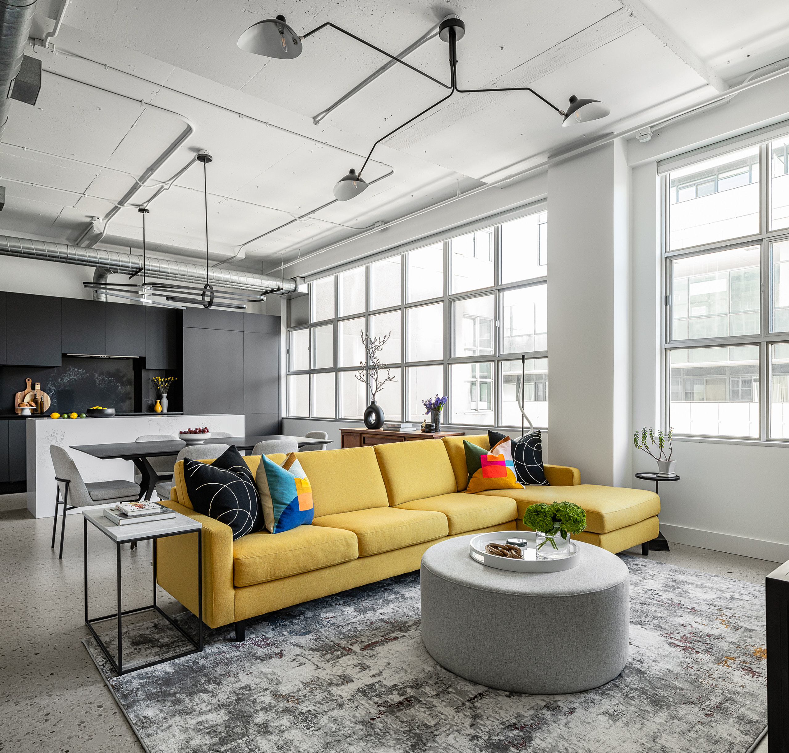 Open concept living room in a converted industrial loft showcasing a bold yellow sectional sofa, concrete epoxy floors, and a black kitchen.