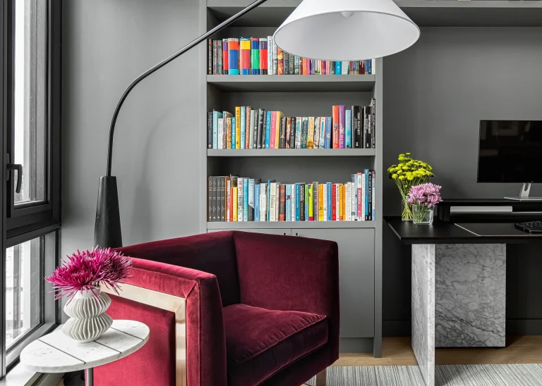 Close-up of a cozy reading corner in the Esplanade home office showcasing a plum velvet chair, modern floor lamp, and custom charcoal bookshelves.