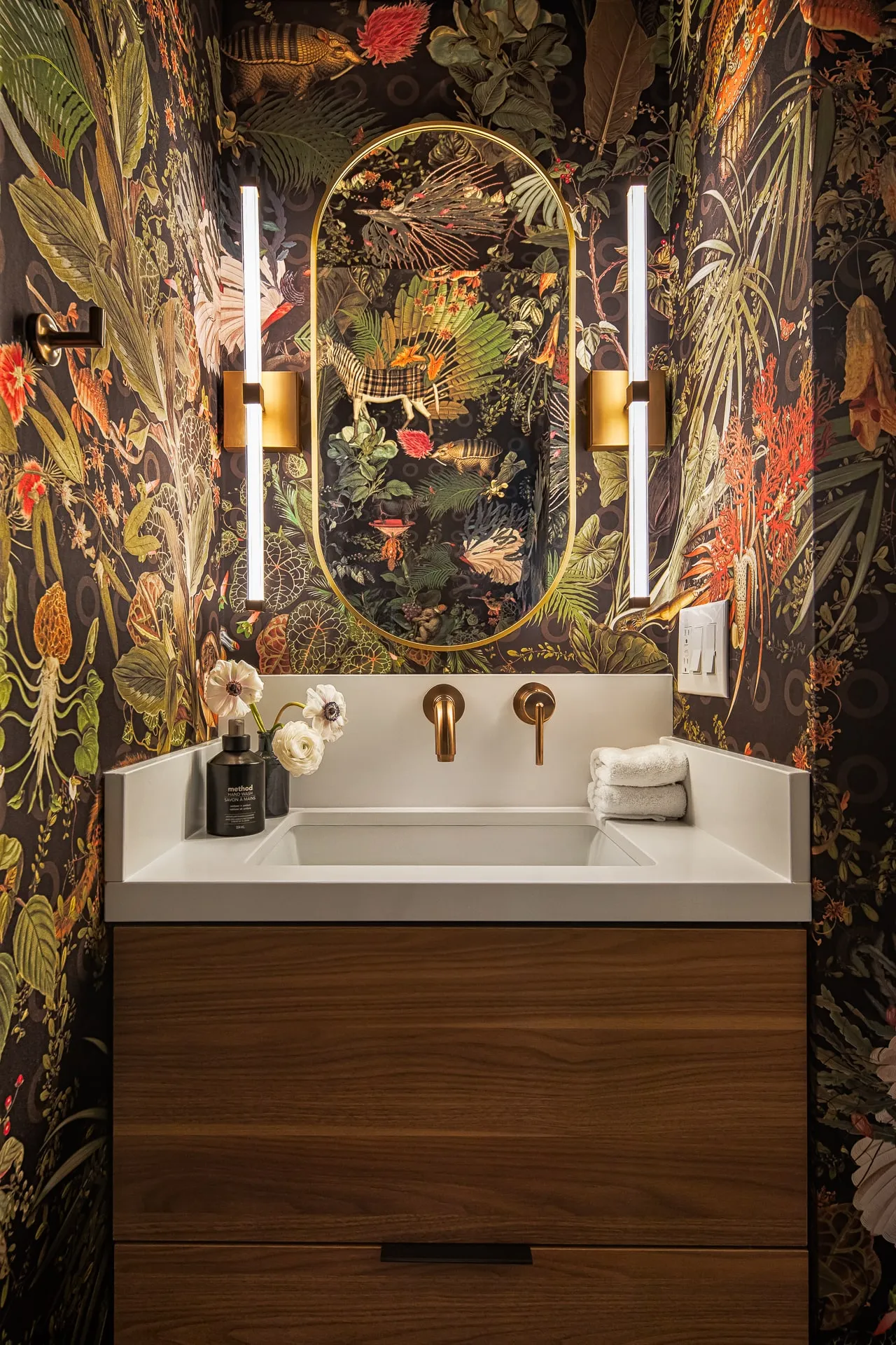 Walnut floating vanity with brass fixtures and oval mirror against dark botanical wallpaper