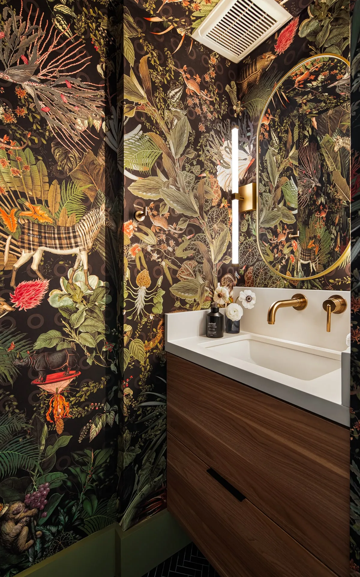 Walnut floating vanity with brass fixtures and oval mirror in a powder room with dark botanical wallpaper