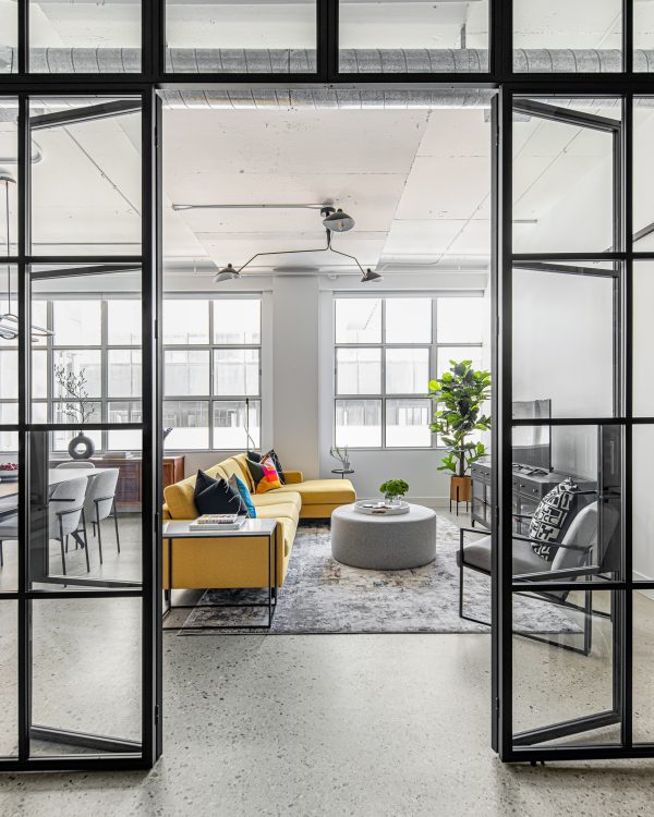 Looking through open custom black metal and glass library doors into a bright loft living room featuring a yellow sectional and exposed concrete ceilings.